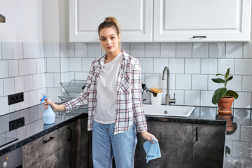 Confident housewife in casual wear with blue rag and detergent in hands. cleaning time in kitchen, looking at camera, posing, at home indoors