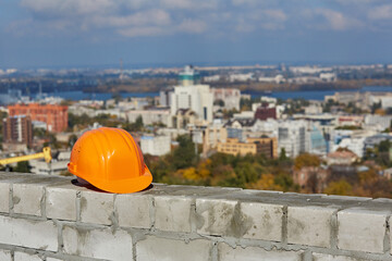 Orange plastic safety helmet is on a brick wall. Roof of modern building under construction. Blurred panoramic cityscape, blue sky, sunny day