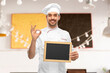 cooking, culinary and people concept - happy smiling male chef in toque showing chalkboard and ok hand sign over restaurant background