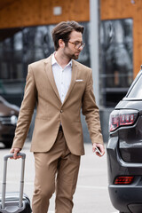 Wall Mural - man in beige suit and glasses holding smartphone and standing near car with suitcase