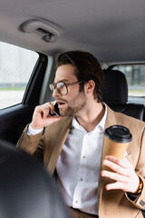 Wall Mural - shocked man in suit and glasses holding paper cup and talking on smartphone in car