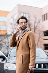 Wall Mural - young man in glasses and coat talking on smartphone outside