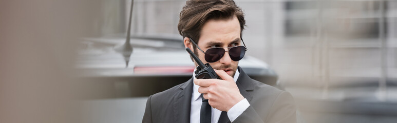 Wall Mural - bodyguard in sunglasses and suit using walkie talkie outside, banner