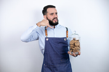 Wall Mural - Young man with beard over isolated white background holding a jar with chocolate chips cookies confused doing phone gesture with hand and fingers like talking on the telephone