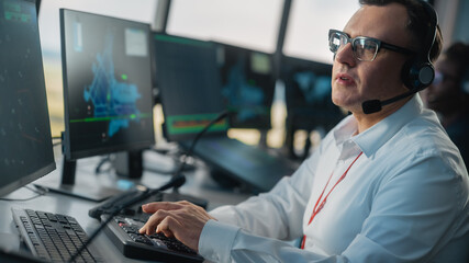 Wall Mural - Male Air Traffic Controller with Headset Talk on a Call in Airport Tower. Office Room is Full of Desktop Computer Displays with Navigation Screens, Airplane Flight Radar Data for the Team. Dutch Angle