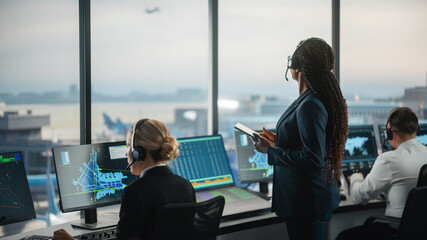 Wall Mural - Black Female Air Traffic Controller Holding Tablet in Airport Tower. Office Room is Full of Desktop Computer Displays with Navigation Screens, Airplane Departure and Arrival Data for the Team.