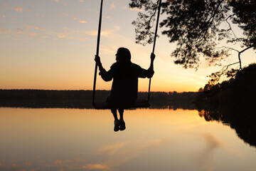 silhouette of a romantic young woman on a swing over lake at sunset. Young girl traveler sitting on the swing in beautiful nature, view on the lake