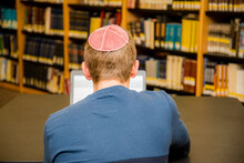 Young Man Wearing A Yarmulke From The Back Doing Work On A Laptop In A Library With Colorful Books On Shelves.
