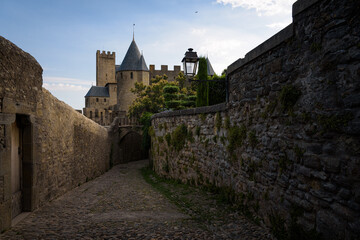 Medieval castle towers stand out in the background over the stone walls of an cobbled alley in the Cite of the fortified city of Carcassonne at sunrise, UNESCO World Heritage Site, France