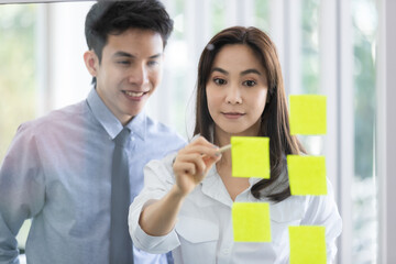 Beautiful black long hair Asian businesswoman and gook looking cute Asian businessman join together in discussion and brainstorming, they write the idea on colorful memo paper on glass board