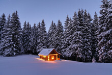 Wooden Hut On The Lawn Covered With Snow. The Lamps Light Up The House At The Evening Time. Winter Landscape. Mystical Night. Mountains And Forests. Wallpaper Background.