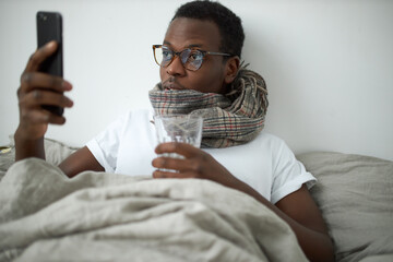 Portrait of sick young dark skinned male suffering from seasonal flu or cold wearing woolen scarf drinking water and browsing internet on smart phone. Unhealthy man working remotely from bed