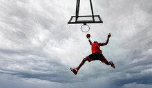 Street Basketball Player Making A Powerful Slam Dunk On The Court - Athletic Male Training Outdoor On A Cloudy Sky Background - Sport And Competition Concept
