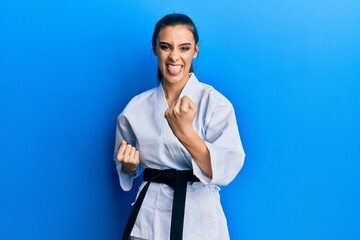 Wall Mural - Beautiful brunette young woman wearing karate fighter uniform with black belt doing attack pose sticking tongue out happy with funny expression.