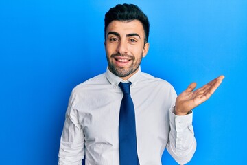 Wall Mural - Young hispanic man wearing business clothes smiling cheerful presenting and pointing with palm of hand looking at the camera.