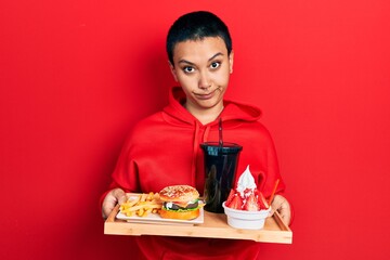 Wall Mural - Beautiful hispanic woman with short hair eating a tasty classic burger with fries and soda skeptic and nervous, frowning upset because of problem. negative person.