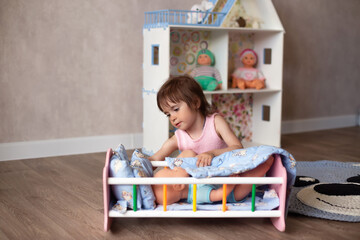 Cute little girl is playing with a doll at home. The child puts the doll to sleep in a toy bed. Role-playing games for children.