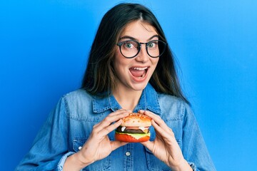 Canvas Print - Young caucasian woman eating a tasty classic burger celebrating crazy and amazed for success with open eyes screaming excited.