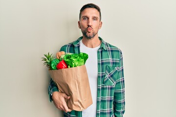 Poster - Handsome man with beard holding paper bag with groceries looking at the camera blowing a kiss on air being lovely and sexy. love expression.