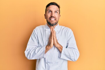 Wall Mural - Handsome man with beard wearing professional cook uniform begging and praying with hands together with hope expression on face very emotional and worried. begging.