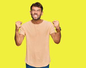 Sticker - Handsome young man with beard wearing casual tshirt angry and mad raising fists frustrated and furious while shouting with anger. rage and aggressive concept.