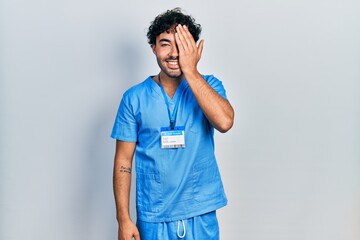 Sticker - Young hispanic man wearing blue male nurse uniform covering one eye with hand, confident smile on face and surprise emotion.