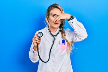 Sticker - Beautiful young blonde doctor woman holding stethoscope peeking in shock covering face and eyes with hand, looking through fingers with embarrassed expression.