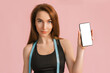 © Daria Lukoiko - Fitness girl smiling and holding a phone with a mockup and posing hold measure tape in black sportswear on a pink background. Slim woman with a beautiful athletic body and tanned skin