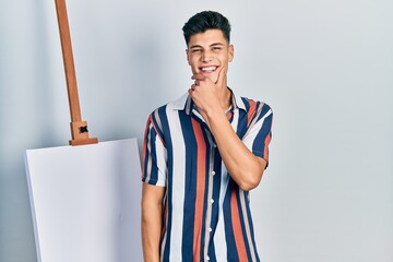 Canvas Print - Young hispanic man standing close to empty canvas looking confident at the camera smiling with crossed arms and hand raised on chin. thinking positive.