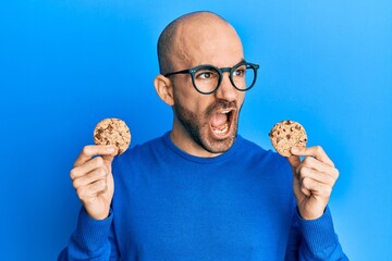 Young hispanic man holding chocolate chips cookies angry and mad screaming frustrated and furious, shouting with anger. rage and aggressive concept.