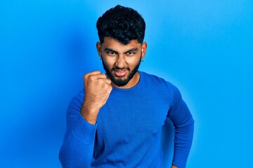 Sticker - Arab man with beard wearing casual blue sweater angry and mad raising fist frustrated and furious while shouting with anger. rage and aggressive concept.