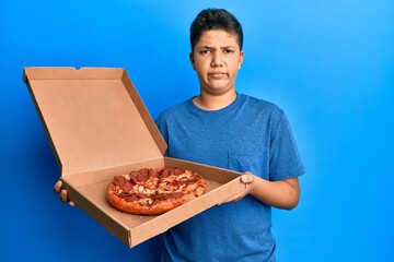 Teenager hispanic boy eating tasty pepperoni pizza skeptic and nervous, frowning upset because of problem. negative person.