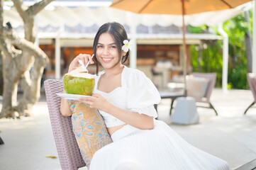 Wall Mural - Beautiful woman tourist with white flower on her hair drinking coconut sitting on lounge chair during summer holidays