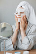 © MyJuly - European woman sitting in front of the mirror and applying a white cloth moisturizing mask on her face, skin care and spa treatments at home, woman beautician makes anti-aging face mask, moisturizing