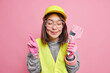 © WHstudio Leushin N - Pleased Asian woman crosses fingers holds paint brush renovates house makes wish believes in good luck wears safety glasses helmet gloves isolated over pink background works at construction company