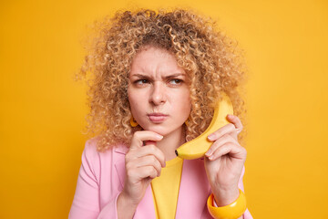 Wall Mural - Serious curly haired European woman with discontent expression pretends having imortant conversation holds tasty banana near ear dressed formally smirks face isolated over yellow background.