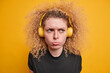 Headshot of displeased curly haired teenage girl has bad mood sulking face expression wears wireless stereo headphones listens music dressed in casual black t shirt isolated over yellow background