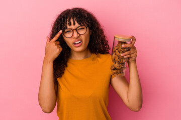 Wall Mural - Young mixed race woman holding a cookies jar isolated on pink background showing a disappointment gesture with forefinger.