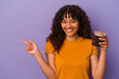 Young mixed race woman holding a coffee beans bottle isolated on purple background smiling and pointing aside, showing something at blank space.