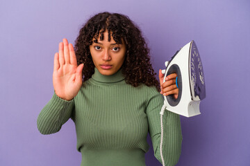 Wall Mural - Young mixed race woman holding an iron isolated on purple background standing with outstretched hand showing stop sign, preventing you.