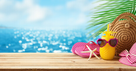 Tropical summer holiday vacation concept with pineapple juice, beach fashion bag and flip flops on wooden table over sea beach background