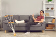 © Studio Romantic - Enjoying recovery sickness benefit after injury in domestic accident. Positive young man with broken leg in plaster cast sitting on sofa at home, using tablet, looking at camera and giving thumbs-up