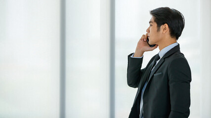 Closeup handsome young Asian businessman wearing formal suit, showing happy expression, smiling with confidence while talking mobile phone, presenting about technology and convenient communication.