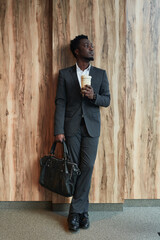 Vertical full length portrait of successful African-American businessman looking away while standing by wood background