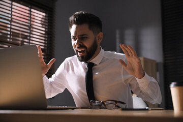 Wall Mural - Emotional young businessman working on laptop in office. Online hate concept