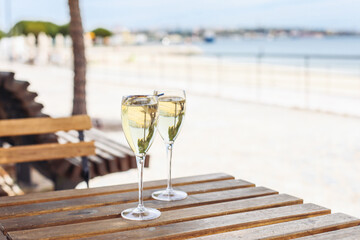 Two glasses of white sparkling wine on a wooden table outdoors. Beach, trees, blue ocean, sunny weather on the background