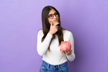 Wall Mural - Young caucasian woman holding a piggybank isolated on purple background having doubts