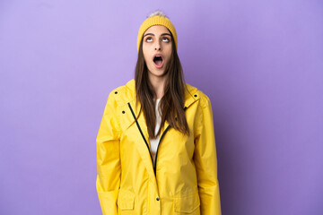 Young caucasian woman wearing a rainproof coat isolated on purple background looking up and with surprised expression