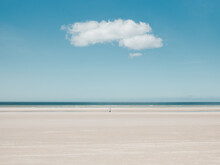 Person Walking Alone Far Away On A Beach With Blue Sky And A Single Cloud