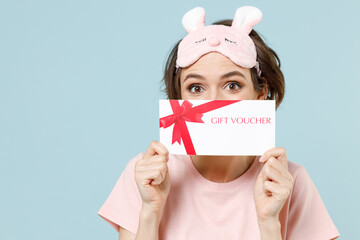 Wall Mural - Young excited woman wear pajamas jam sleep eye mask rest relaxing at home hiding cover face with gift voucher flyer mock up isolated on pastel blue background studio. Good mood night bedtime concept.